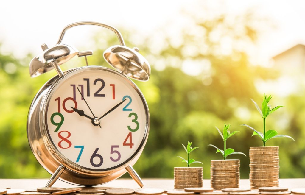 An old fashion alarm clock to the left of three stacks of coins, the first being the shortest and the last being the tallest and each stack of coins has a plant growing out of the top of it