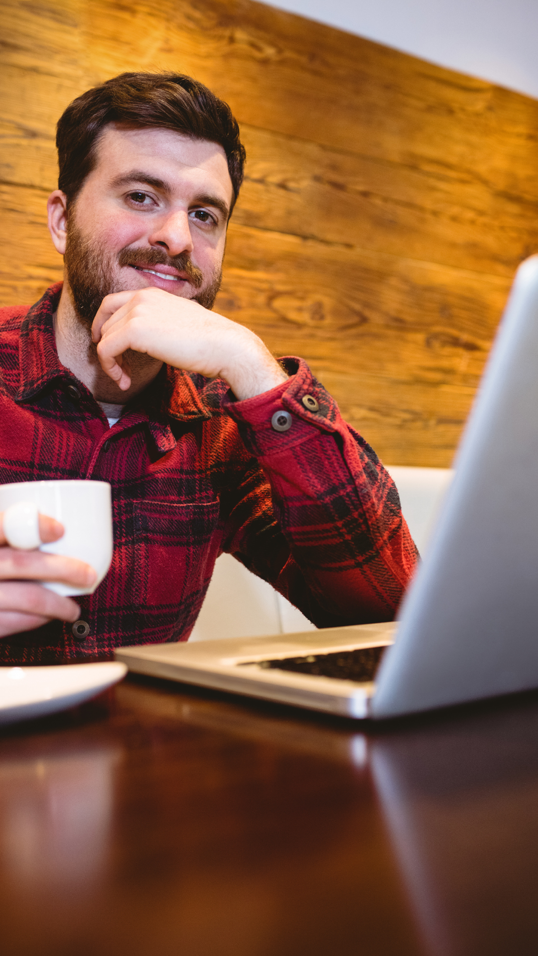 Dark haired, bearded man in red and black flannel shirt smiles as he lifts a white cup of coffee with his right hand, holds his chin with his left hand, left elbow and laptop sit on wood table in front of him