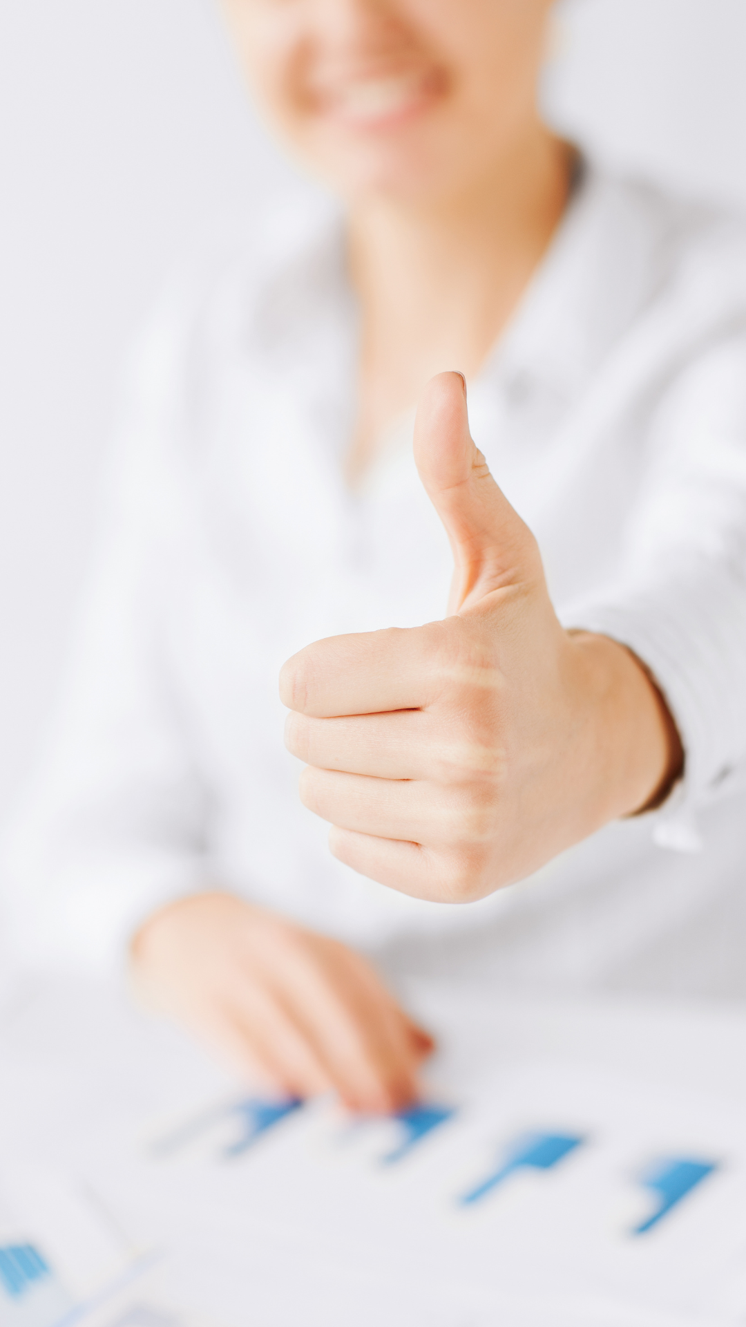 Woman smiling, wearing a white button down shirt sits with graphs on the table in front of her, all blurred, gives a thumbs up in the forefront of the frame